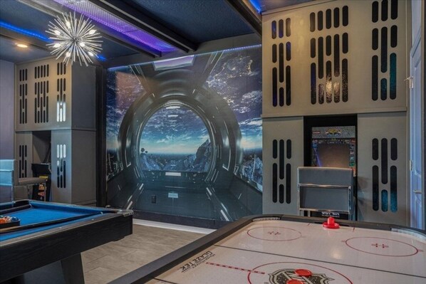 A pool table with a blue felt surface, a shuffleboard table with red and white markings, and a wall with a space-themed mural.