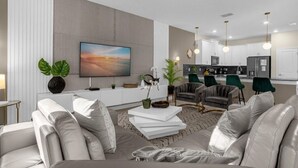 Moderni olohuone, jossa on sohva, sohvapöytä ja seinälle asennettu televisio. Taustalla näkyy tuoleja ja keittiöalue.