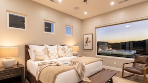 A bedroom with a large bed, bedside lamps, a nightstand, a wooden bench, a leather chair, and a large window with a view.