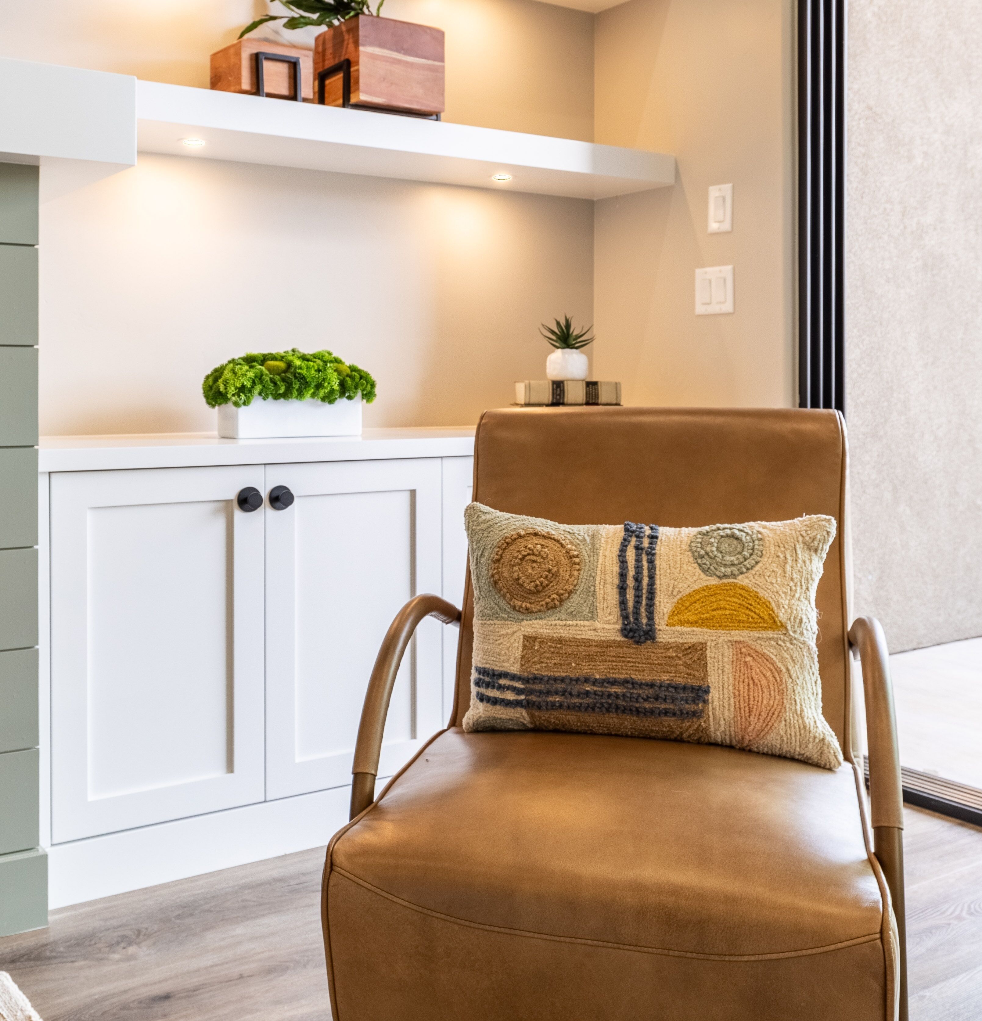 A brown leather chair with a decorative pillow, a white cabinet with a plant, and a shelf with decorative items.