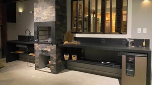A modern kitchen with a built-in stone fireplace, a stainless steel refrigerator, and a large window with blinds. - CASA BALI - Charm and comfort in Guarajuba  (Guarajuba)