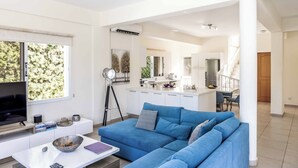 Ein modernes Wohnzimmer mit einem blauen Sofa, einem weißen Couchtisch und einem Fernseher. Im Hintergrund ist eine Küche mit weißen Schränken und einem Essbereich zu sehen.