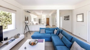 Ein modernes Wohnzimmer mit einem blauen Ecksofa, einem weißen Couchtisch und einem Fernseher auf einem Schrank. Im Hintergrund steht ein Stativ mit einer Kamera, und durch eine offene Tür ist eine Küchenzeile zu sehen.