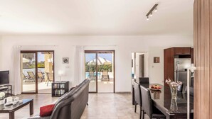 Una sala de estar moderna con un sofá, una mesa de centro y un comedor con una mesa y sillas. Hay un televisor, una estantería y una cocina con un refrigerador.