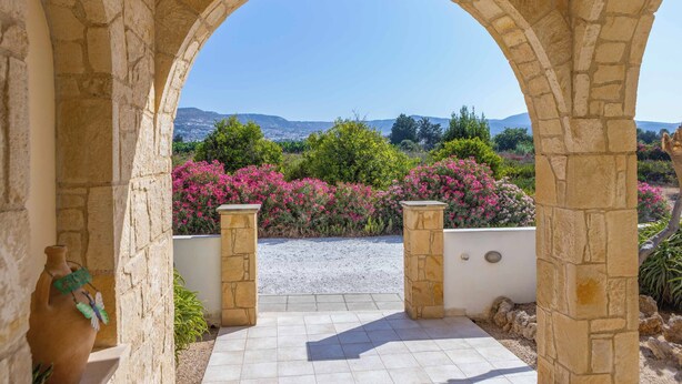 Un arcoado portal de piedra que da a un camino de grava con vistas a una ladera y arbustos en flor.