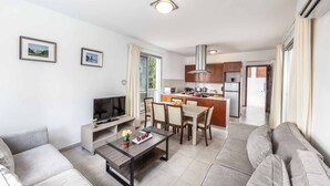 Un salon moderne avec un canapé, une table basse et un téléviseur. On y trouve également un coin repas avec une table et des chaises. La cuisine est visible au fond, avec des placards et une cuisinière.