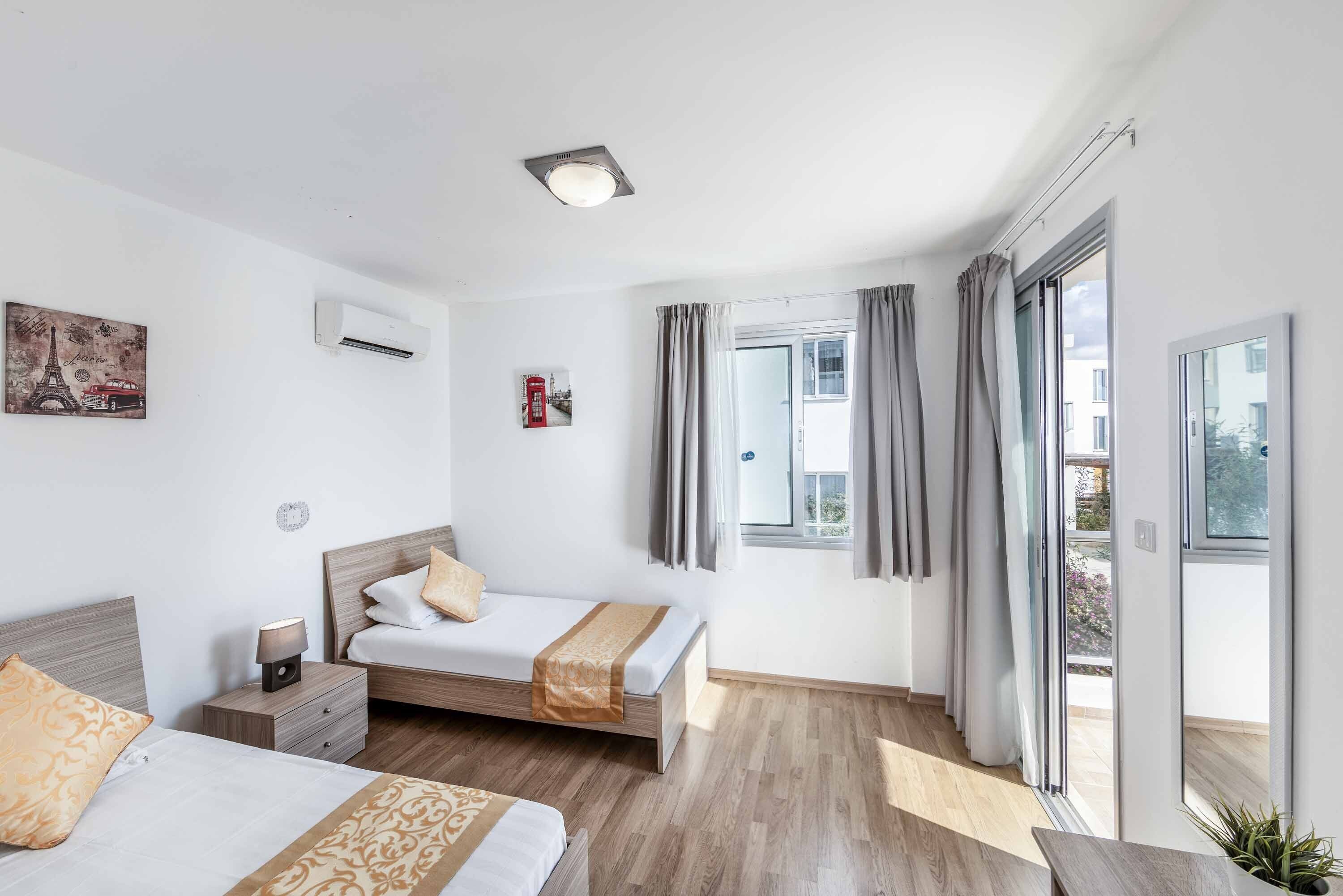 Ein modernes Hotelzimmer mit zwei Betten, einem Nachttisch mit Lampe, einer Wand-Klimaanlage, einem Fenster mit Vorhängen und Blick ins Freie.