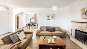 A living room with a sofa, coffee table, fireplace, and dining area in the background. - Coral Olympus Poseidon (Peyia)