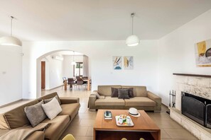 A living room with a sofa, coffee table, fireplace, and dining area in the background. - Coral Olympus Poseidon (Peyia)