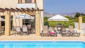 A poolside area with lounge chairs and umbrellas, a table with chairs, and a view of a cityscape in the background.