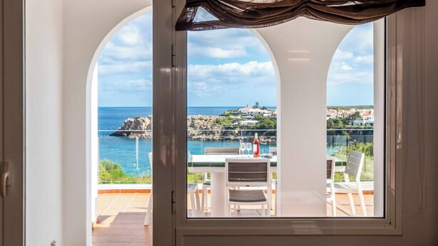 Un balcón con mesa y sillas, con vista al mar y casas a la orilla.
