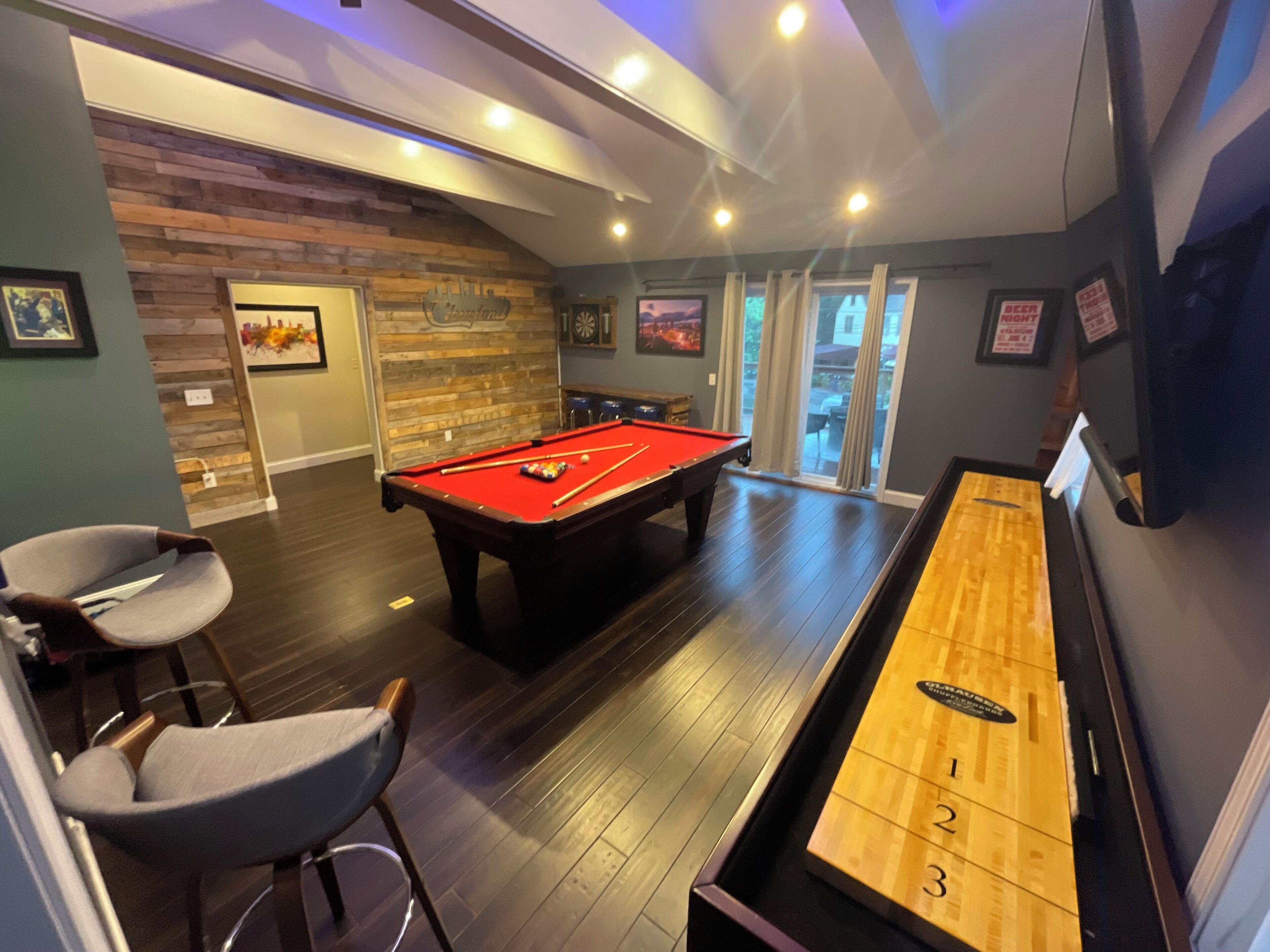 A pool table with a red felt surface, a shuffleboard table, and a billiards table in a room with wooden flooring and a sloped ceiling.