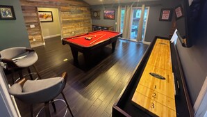Une table de billard recouverte d’un feutre rouge, une table de jeu de palet et une table de billard se trouvent dans une pièce dont le sol est en bois et dont le plafond est incliné.