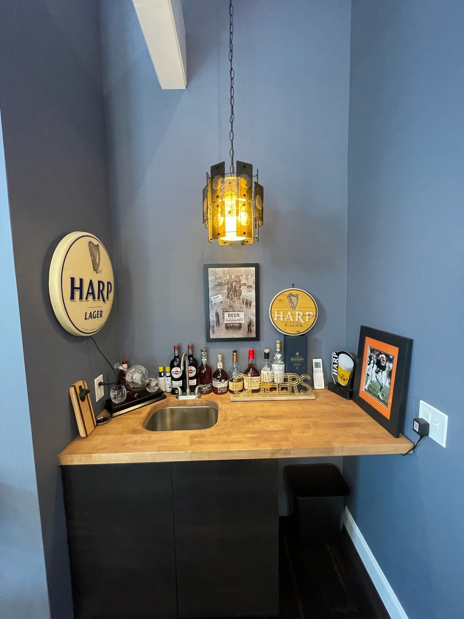 A bar area with a wooden countertop, a sink, and a hanging light fixture. There are several beer taps and bottles on the counter.