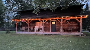Une cabane rustique en rondins de bois, dotée d’un porche, de chaises en bois et d’une porte noire.