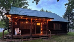 Une cabane rustique en rondins de bois, dotée d’un porche, d’un banc en bois et de guirlandes lumineuses.