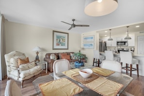 A modern living room with a glass dining table, a sofa, and a ceiling fan. - 1BR Coastal Condo + 3 Pools & Private Beach Access | Magnolia House 107 (Destin)