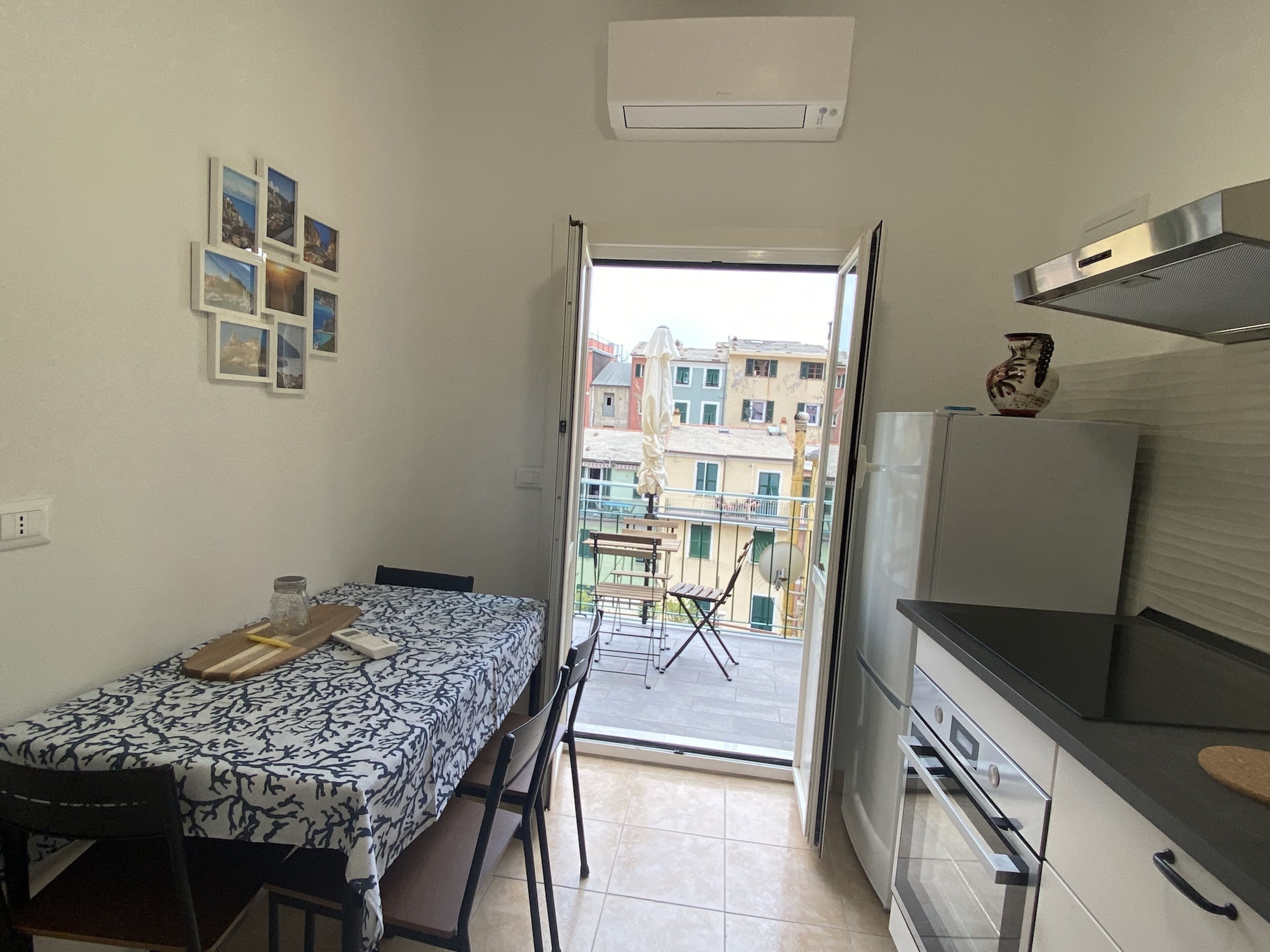 A compact kitchen with a dining area, a view of the city through an open door, and a small balcony with a chair and umbrella.