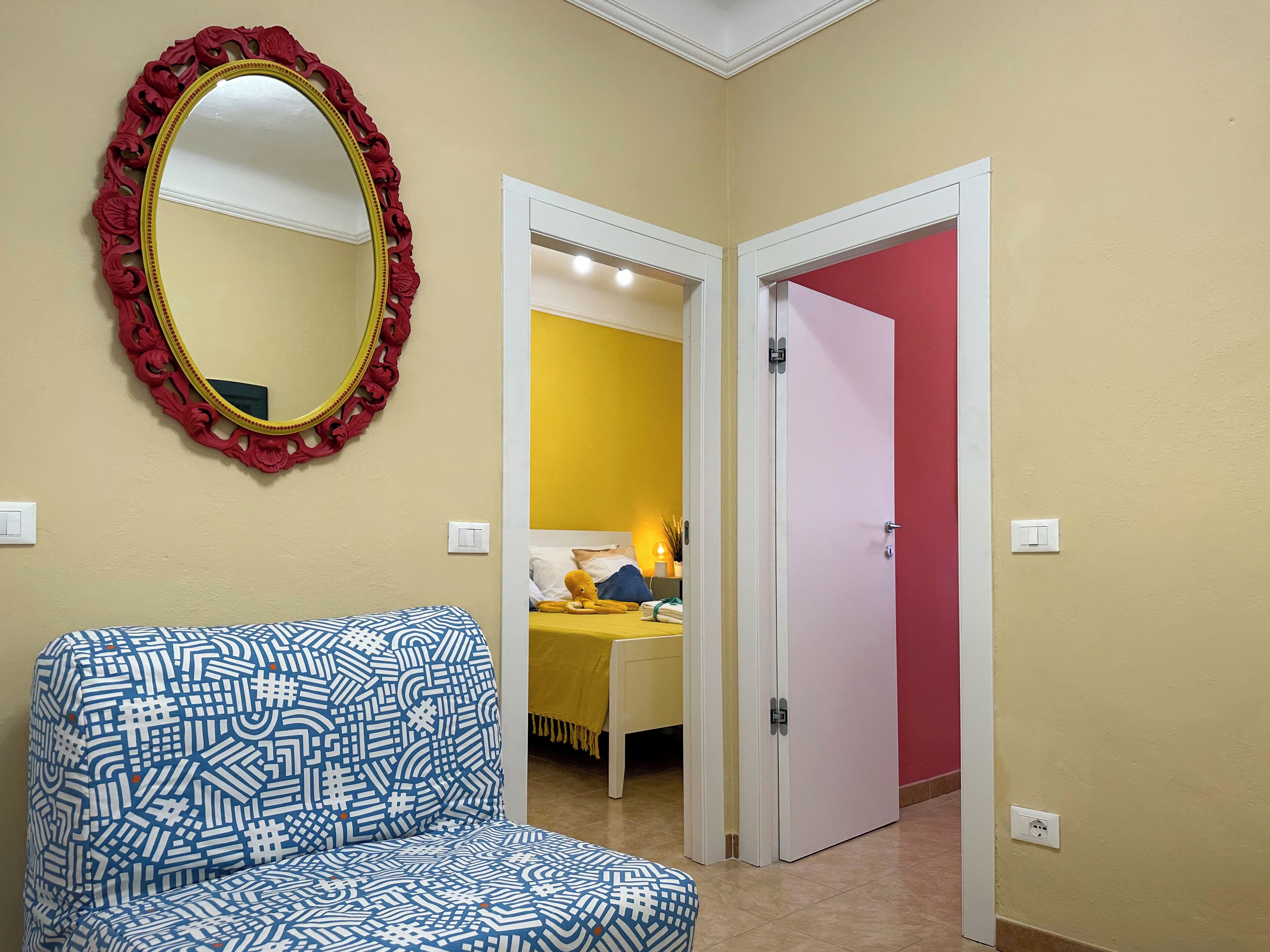 Une pièce avec une chaise à motifs, un miroir avec un cadre rouge et un lit avec une literie jaune.