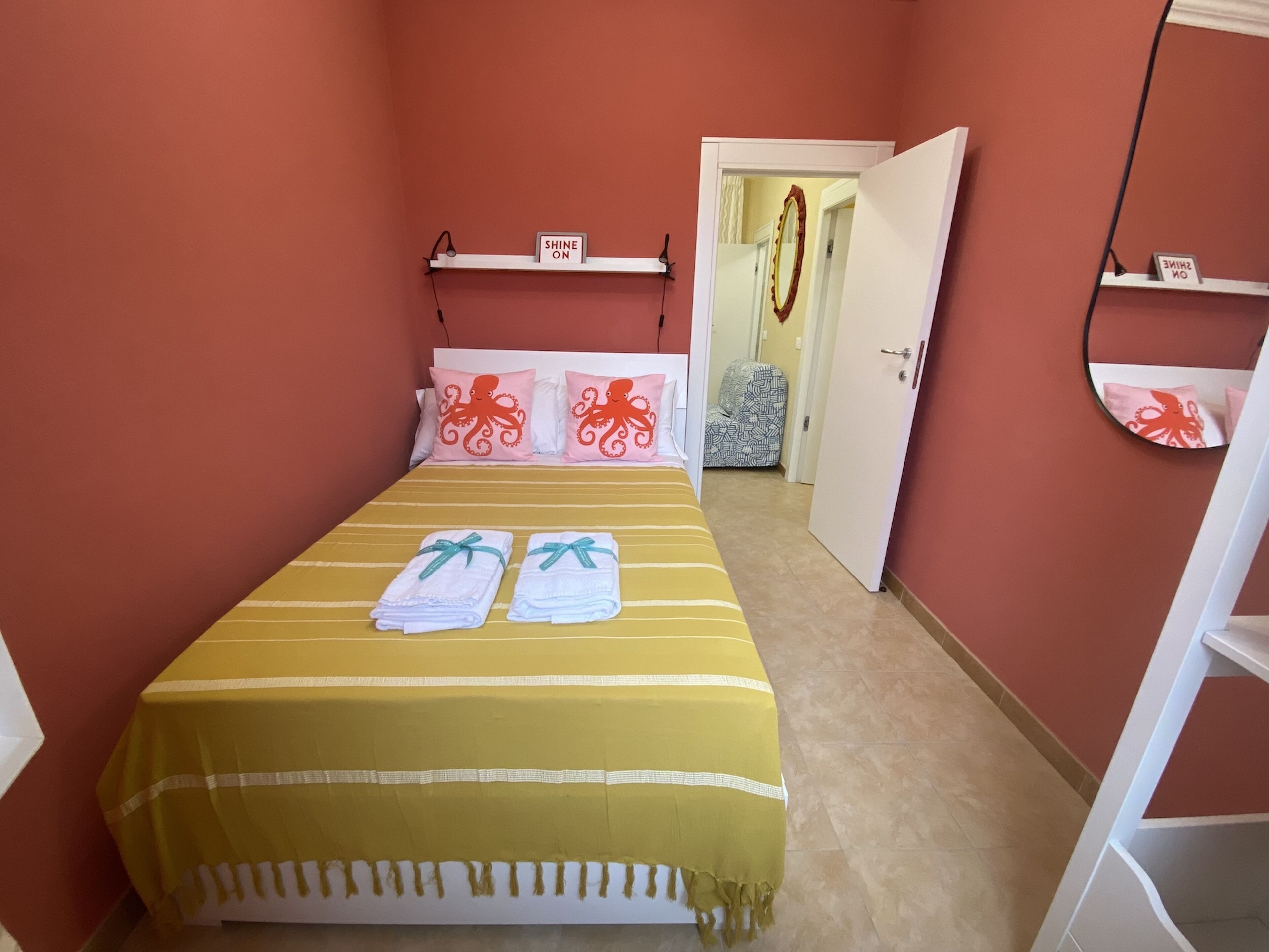 Una habitación con una cama, un espejo, una estantería con un cartel y una puerta que da a otra estancia.