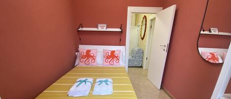 Una habitación con una cama, un espejo, una estantería con un cartel y una puerta que da a otra estancia.