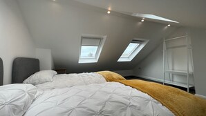 A bedroom with a sloped ceiling, two skylights, a bed with white bedding, a yellow throw, and a white shelf unit. - Sea Haven  Stylish Beach Oasis - Pass the Keys (Hastings)