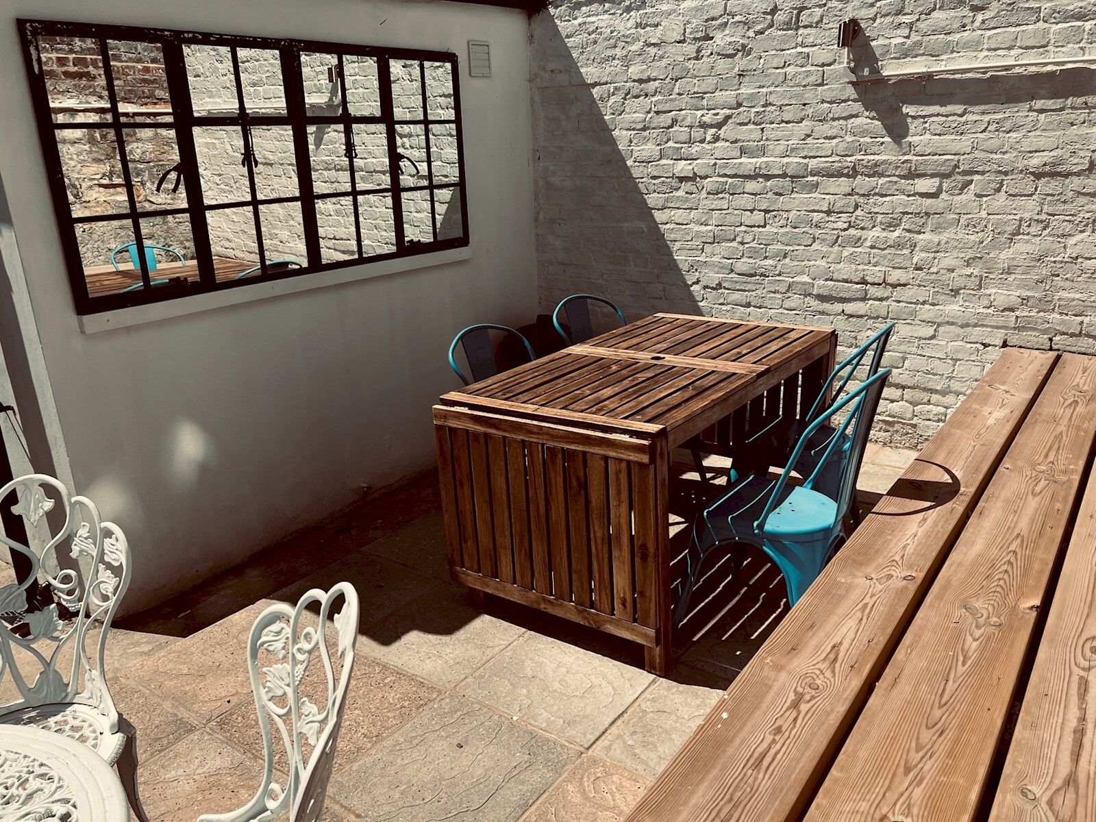 Eine Terrasse mit Holztisch und -stühlen, eine weiße Wand mit Fenster und ein Backsteinwall im Hintergrund.