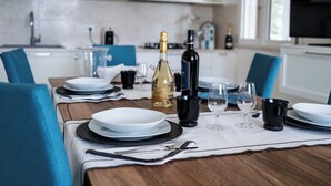 A dining table set with plates, glasses, and a bottle of wine in a modern kitchen with white cabinets and a TV in the background.