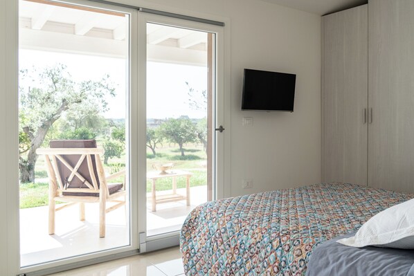 Un dormitorio con cama, televisor montado en la pared y puerta corrediza de vidrio que da a un espacio exterior con una silla y una mesa.