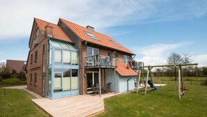 A two-story house with a large glass window, a balcony, and a wooden deck. - 18 Ferienwohnung im Fritz-bleyl-weg 16a EG (Burg auf Fehmarn)