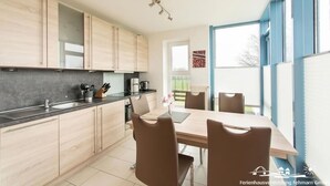 A modern kitchen with light wood cabinets, a black countertop, and a dining area with a wooden table and brown chairs. - 18 Ferienwohnung im Fritz-bleyl-weg 16a EG (Burg auf Fehmarn)