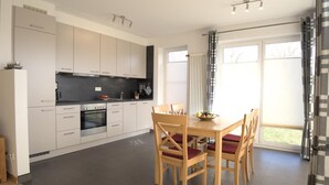 Une cuisine moderne avec des meubles blancs, une table à manger entourée de quatre chaises et une vue sur l’extérieur grâce à une grande fenêtre.