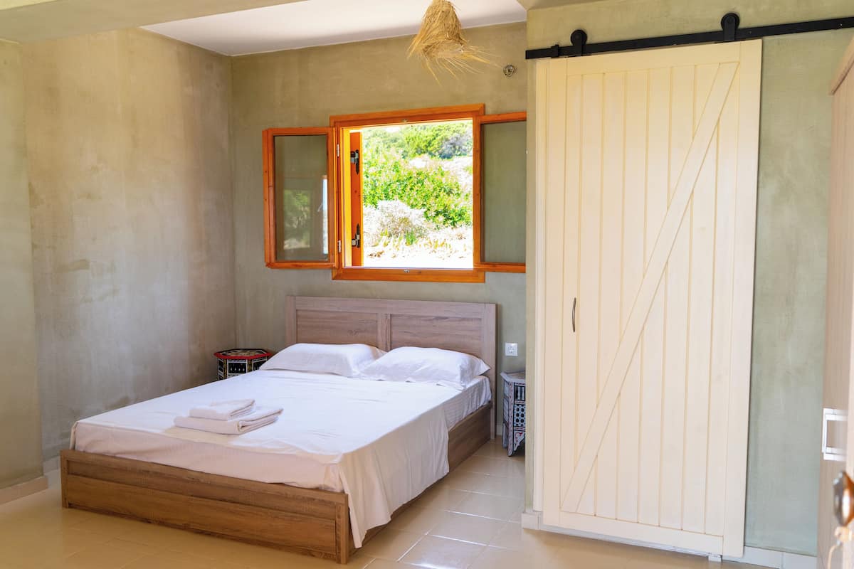 A bedroom with a wooden bed, a nightstand, a wardrobe, and a window with a view of greenery.