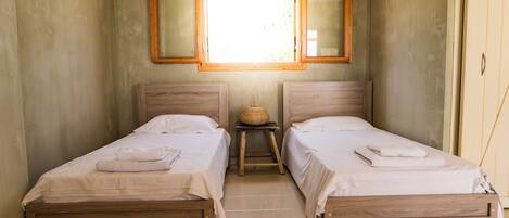 Una habitación pequeña y limpia con dos camas de madera, un taburete pequeño y una ventana con marcos de madera.