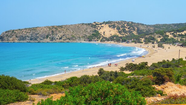 Une vue côtière avec une plage de sable, une mer bleue et limpide, et des collines verdoyantes en arrière-plan.