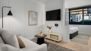 Ein modernes Hotelzimmer mit Sofa, kleinem runden Tisch, Fernseher, Nachttisch und Fenster mit Blick auf Gebäude.