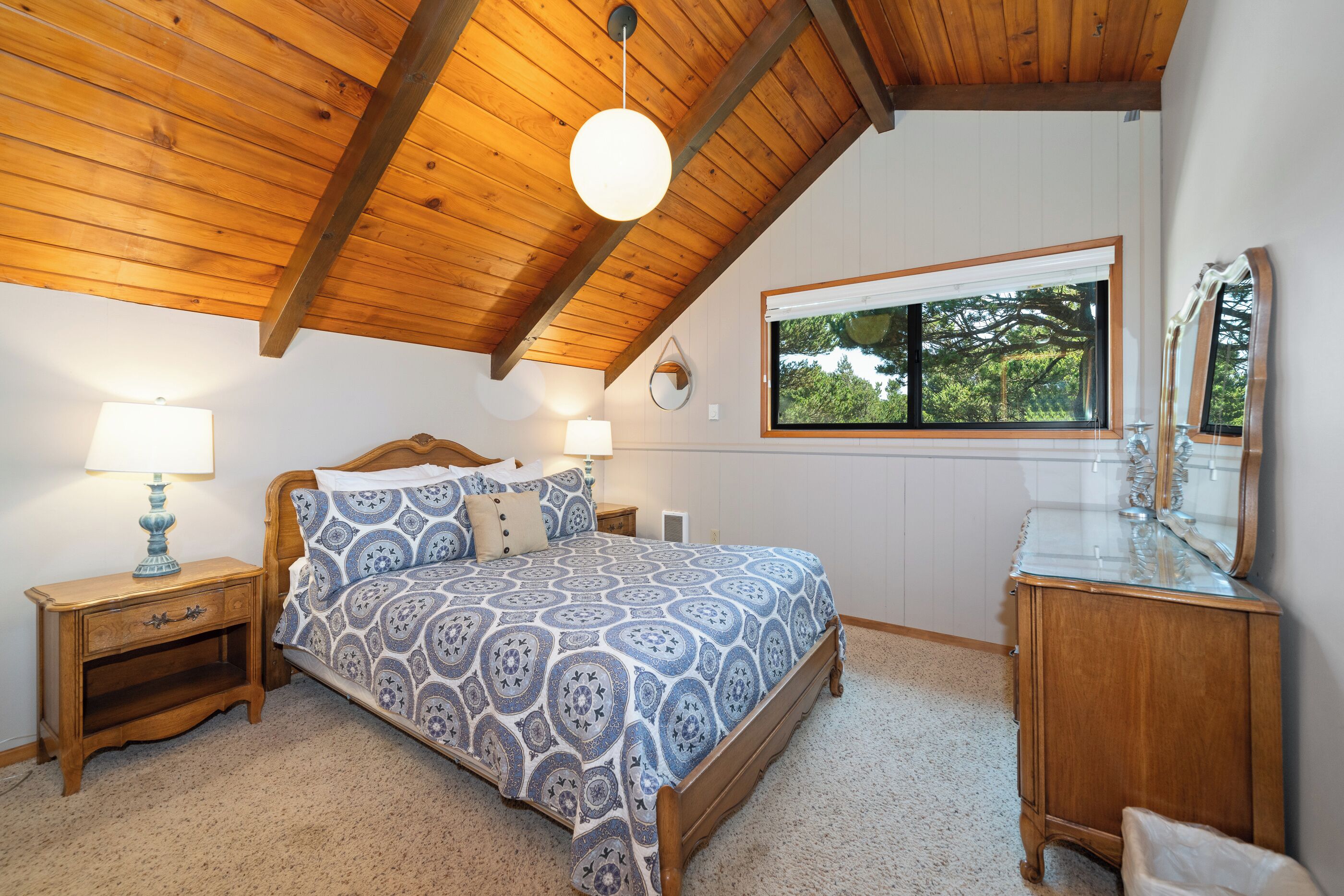 A bedroom with a wooden ceiling, a bed with patterned bedding, a nightstand with a lamp, a dresser with a mirror, and a window with a view of trees.