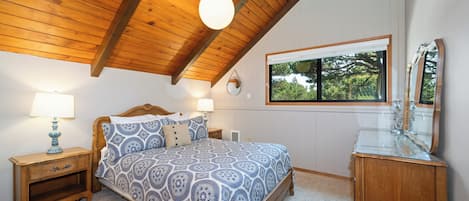 A bedroom with a wooden ceiling, a bed with patterned bedding, a nightstand with a lamp, a dresser with a mirror, and a window with a view of trees.