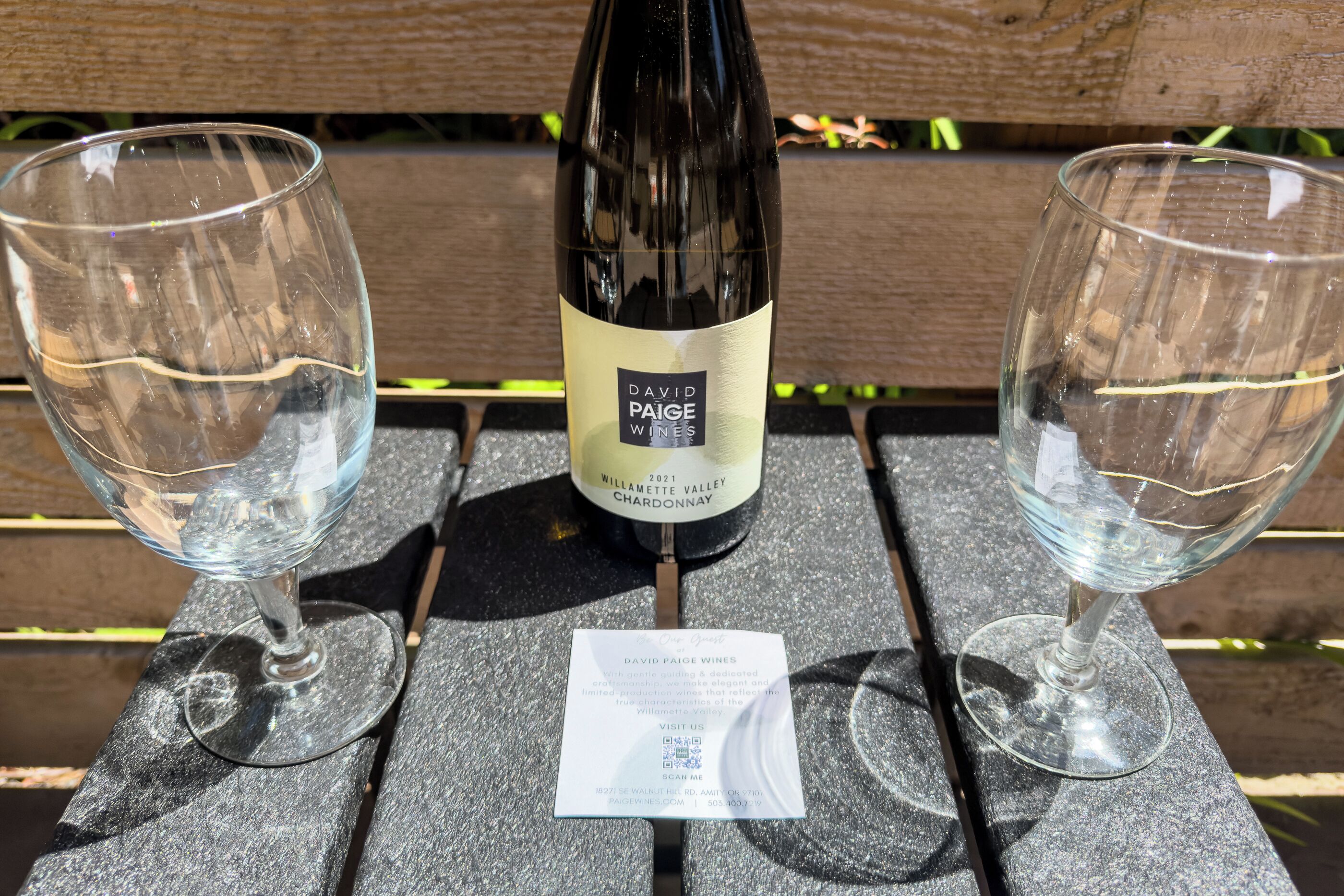 A bottle of David Paige Wines 2021 Willamette Valley Chardonnay on a table with two empty wine glasses.
