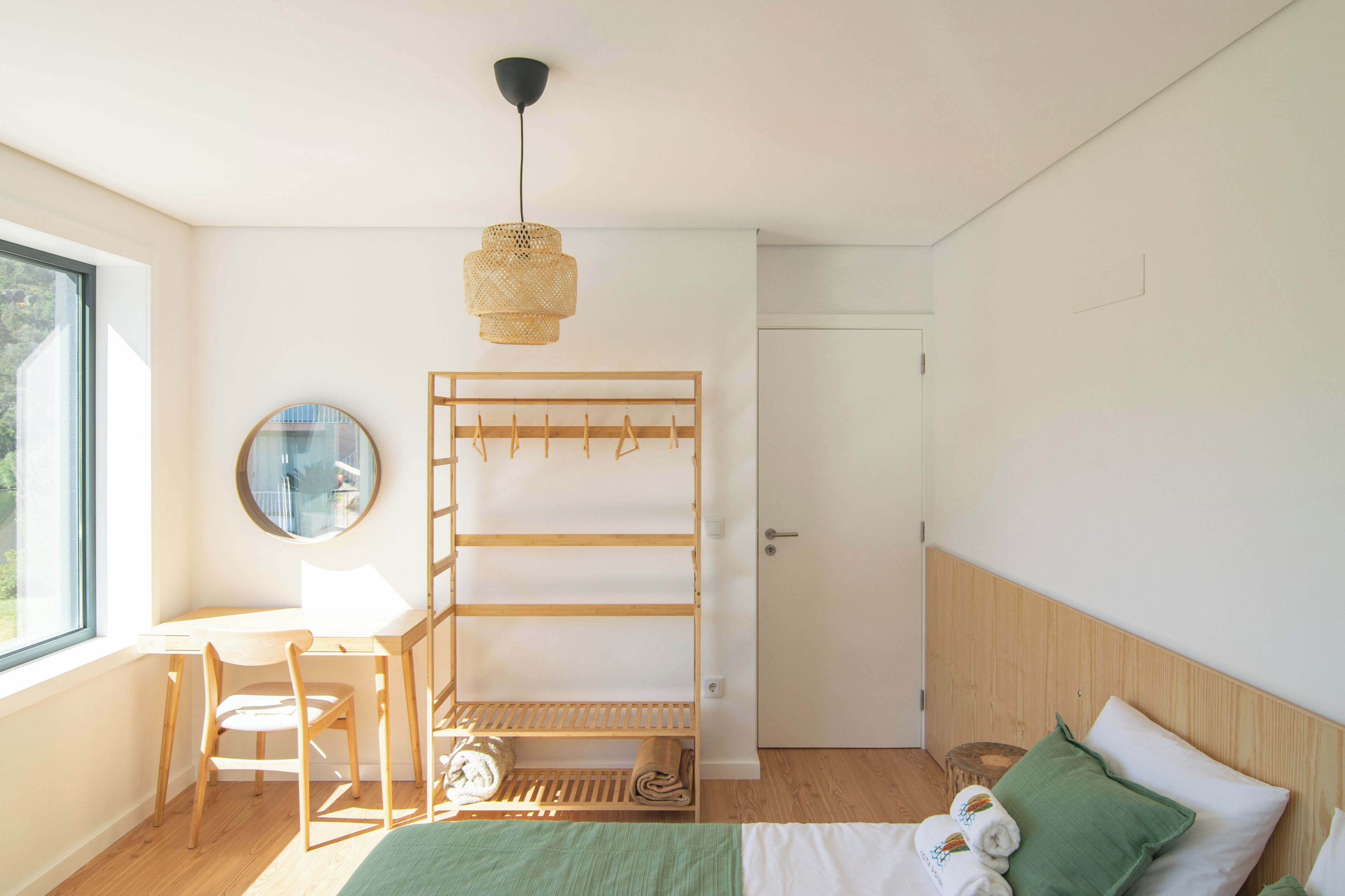 A bedroom with a bed, a desk, a chair, a hanging rack, a mirror, and a window.