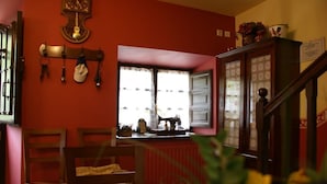A room with a red wall, a wooden table, two chairs, a window with a view, and a cabinet with a mirror. - Green House (Fuentes)