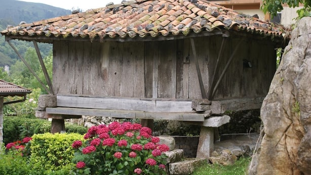 A rustic wooden structure with a tiled roof, surrounded by lush greenery and flowering plants. - Green House (Fuentes)