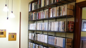 A bookshelf filled with books in a room with a red and yellow wall, a wooden floor, and a small table with a chair. - Green House (Fuentes)