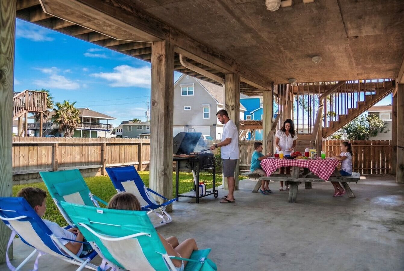 A covered outdoor area with lounge chairs, a barbecue grill, and a picnic table with a checkered tablecloth.