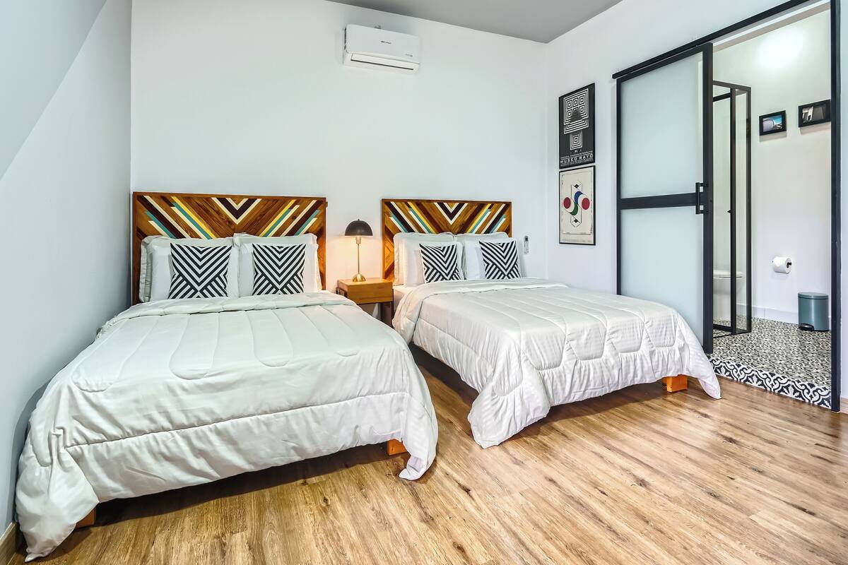 Habitación con dos camas, piso de madera, aire acondicionado instalado en la pared y puerta corredera de vidrio hacia el baño.