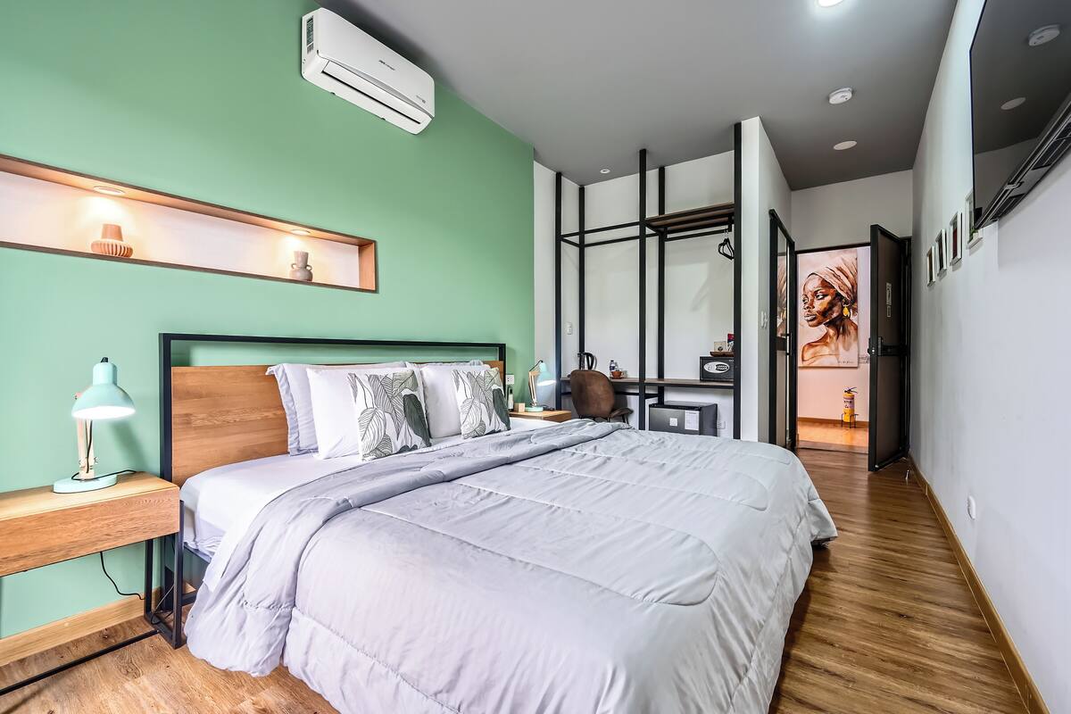 Un dormitorio moderno con una cama grande, un escritorio y una silla. Hay una estantería con objetos decorativos y un equipo de aire acondicionado en la pared.
