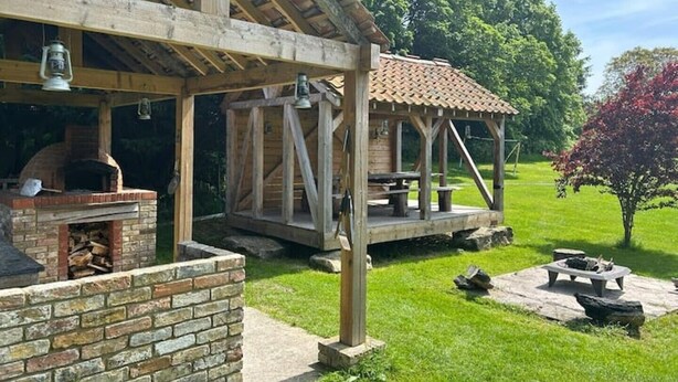A rustic outdoor patio with a brick oven, wooden pergola, and picnic table. - Charming Group Stay – Peaceful Retreat near London (Bexley,)