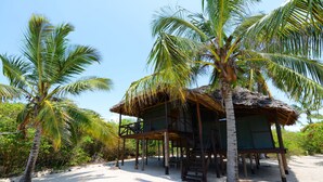 A beachside hut with a thatched roof, surrounded by palm trees and sandy ground. - Simply Saadani Camp (Mkwaja)