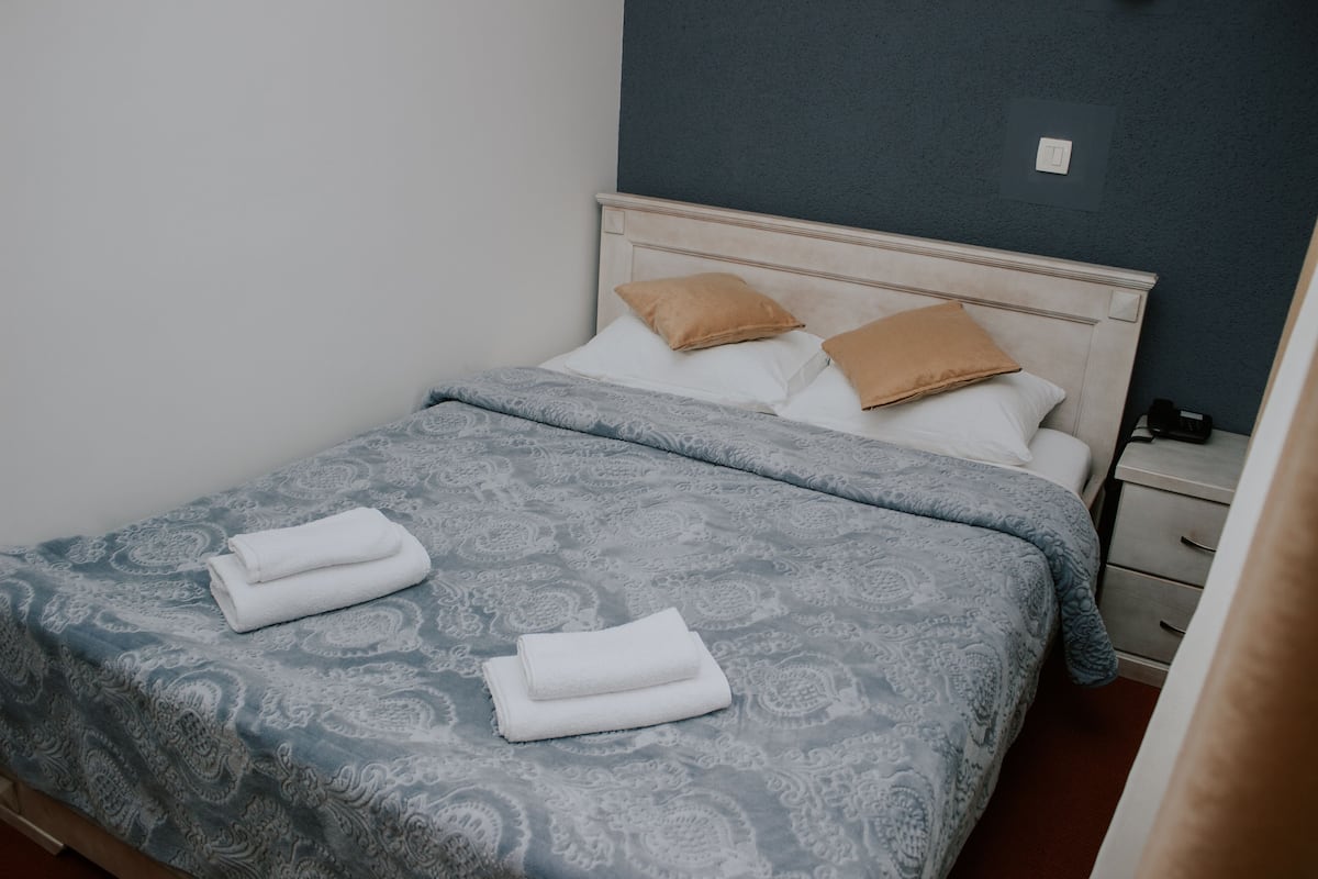 Cama bem arrumada com colcha estampada e duas almofadas, mesa de cabeceira com abajur e interruptor de parede.