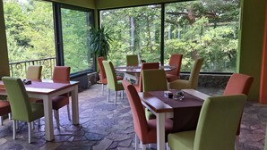 Un espacio de comedor al aire libre, cubierto, con piso de piedra y ventanas amplias que dan vista a un área boscosa.